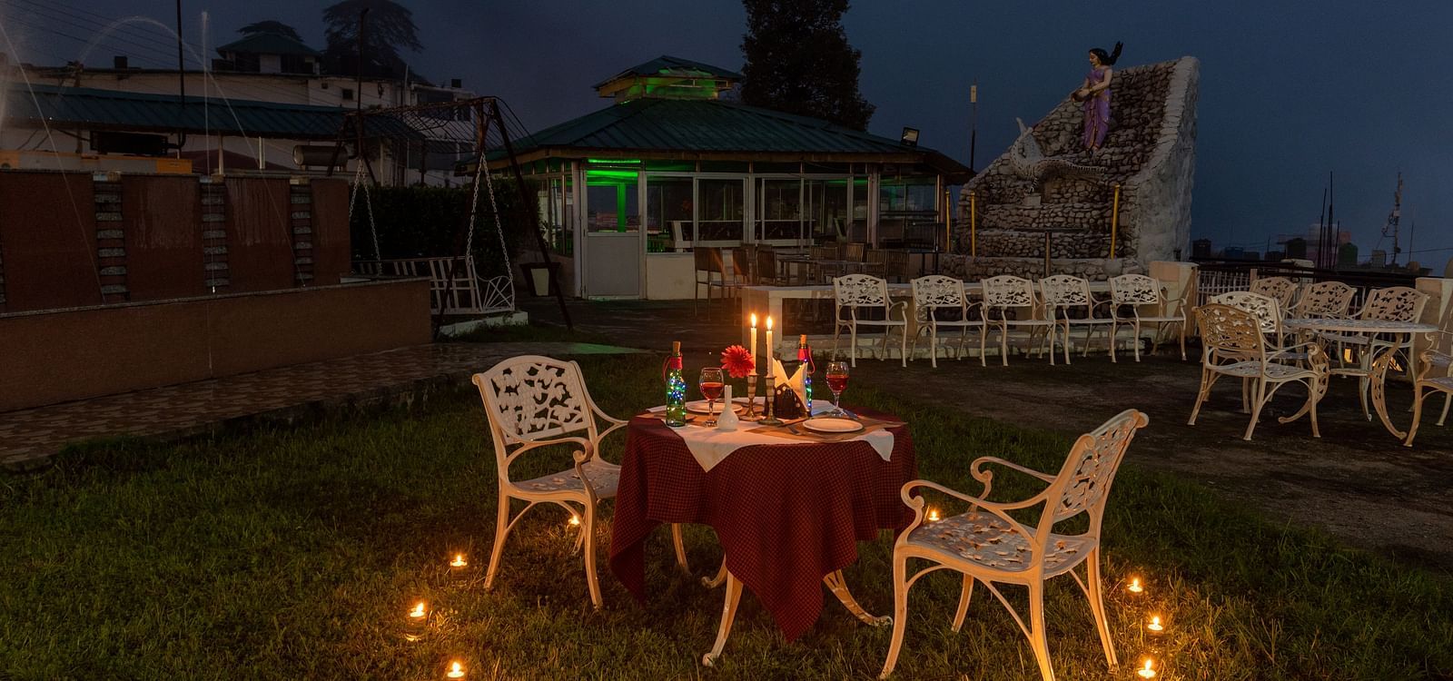 Candlelight dinner setup with a table for two, surrounded by candles and a view of a small fountain and seating area in the background - DLS Meghavan Resort, Dharamshala